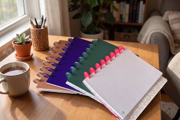 Carnets Mylibup en éventail sur un bureau en bois près d'une fenêtre, avec un carnet ouvert sur une recharge à lignes, une tasse de thé et une plante