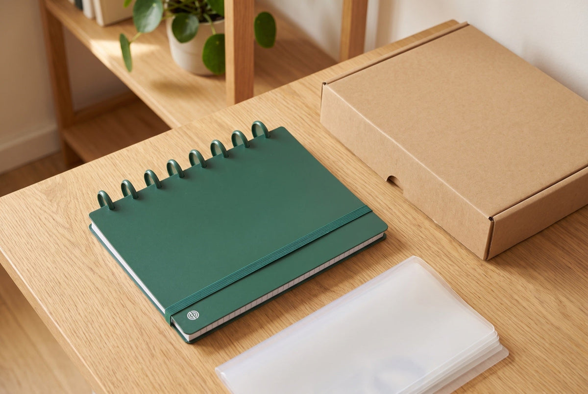 Carnet Mylibup vert fermé sur un bureau en bois clair, avec une boîte d'archives en carton kraft et des pochettes transparentes