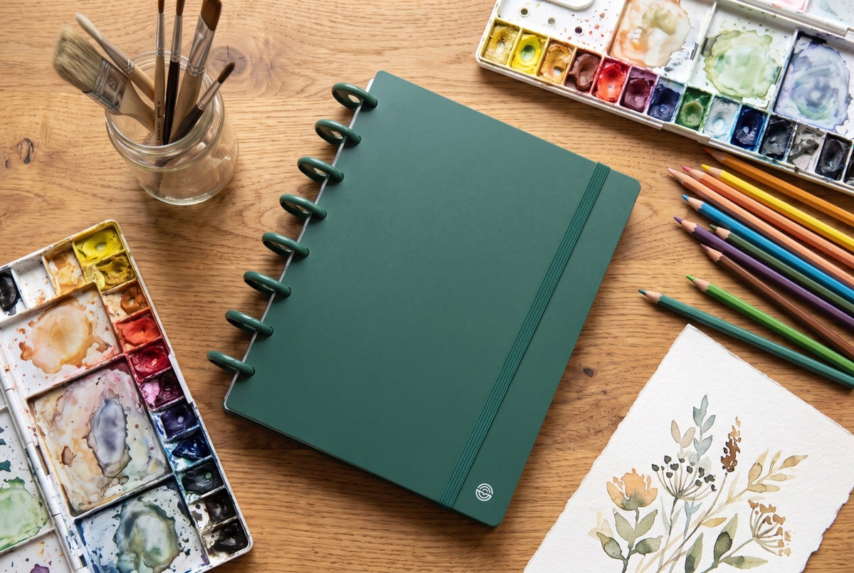 Carnet Mylibup vert posé sur une table en bois, entouré de pinceaux, d'une palette d'aquarelle, de crayons de couleur et d'une aquarelle florale