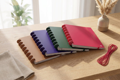 Quatre carnets Mylibup en éventail (camel, violet, vert, framboise) sur une table en bois près d'une fenêtre, avec un vase de fleurs séchées et un élastique