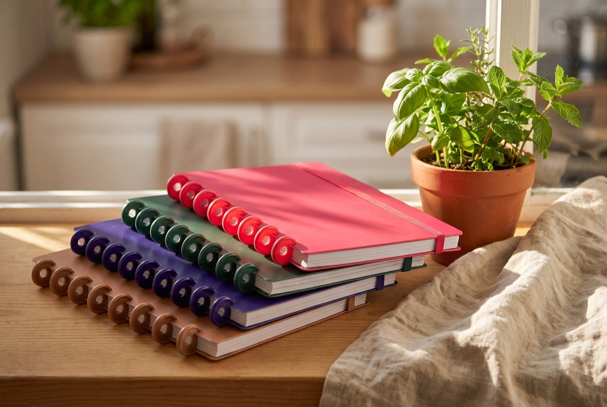 Carnets Mylibup empilés (camel, violet, vert, framboise) sur un rebord de fenêtre ensoleillé, avec un pot d'herbes fraîches et un torchon en lin